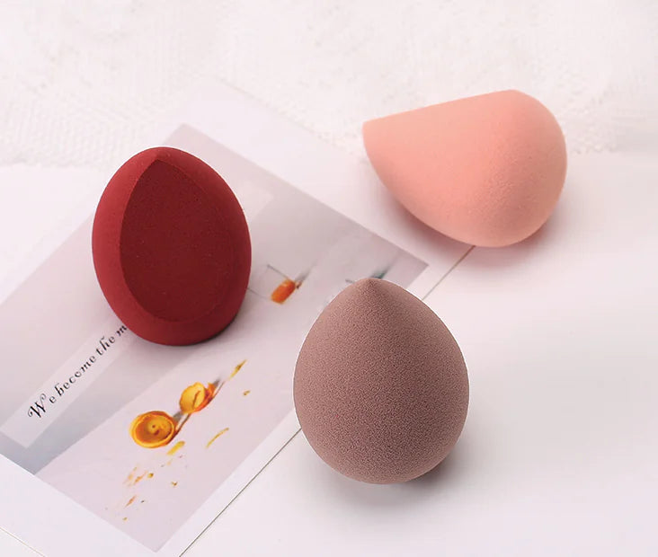Three makeup sponges in red, pink, and brown on a white surface with a magazine underneath.