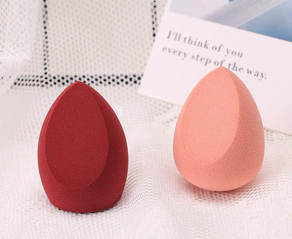 Two makeup sponges, one red and one pink, on a textured white surface.