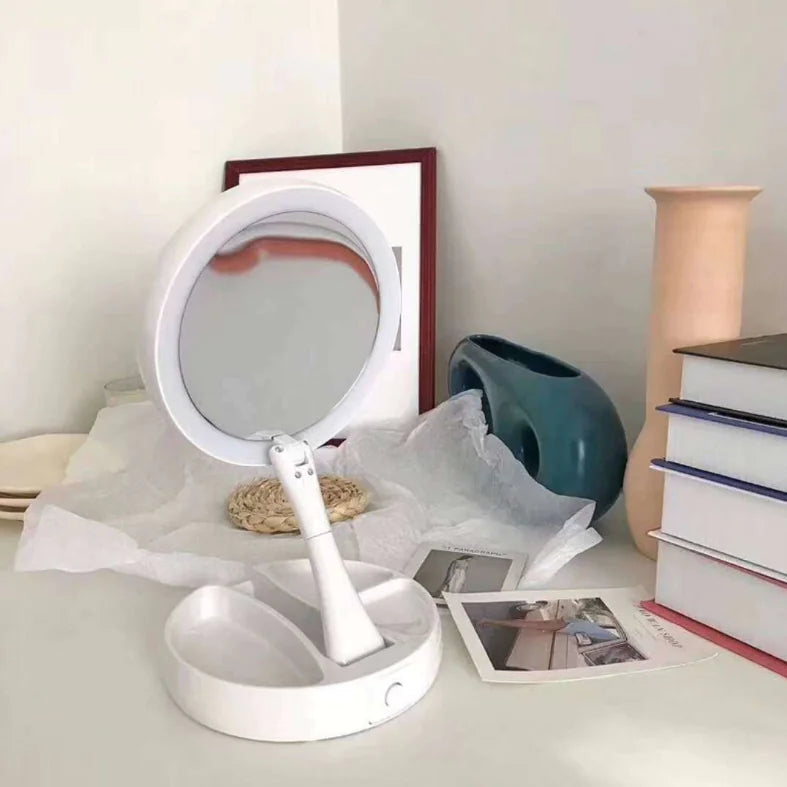 Round white makeup mirror with a stand on a surface with books and a vase.